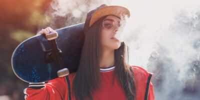 Vaping girl. Young hipster woman vape e-cig. Pretty young female in black hat, red clothing vape ecig, vaping device at the sunset. Toned image. Closeup. Hip-hop style.